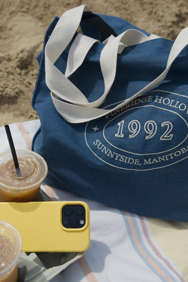 Blue tote bag with '1992 Sunnyside, Manitoba' text on a sandy surface with a yellow phone and drinks.
