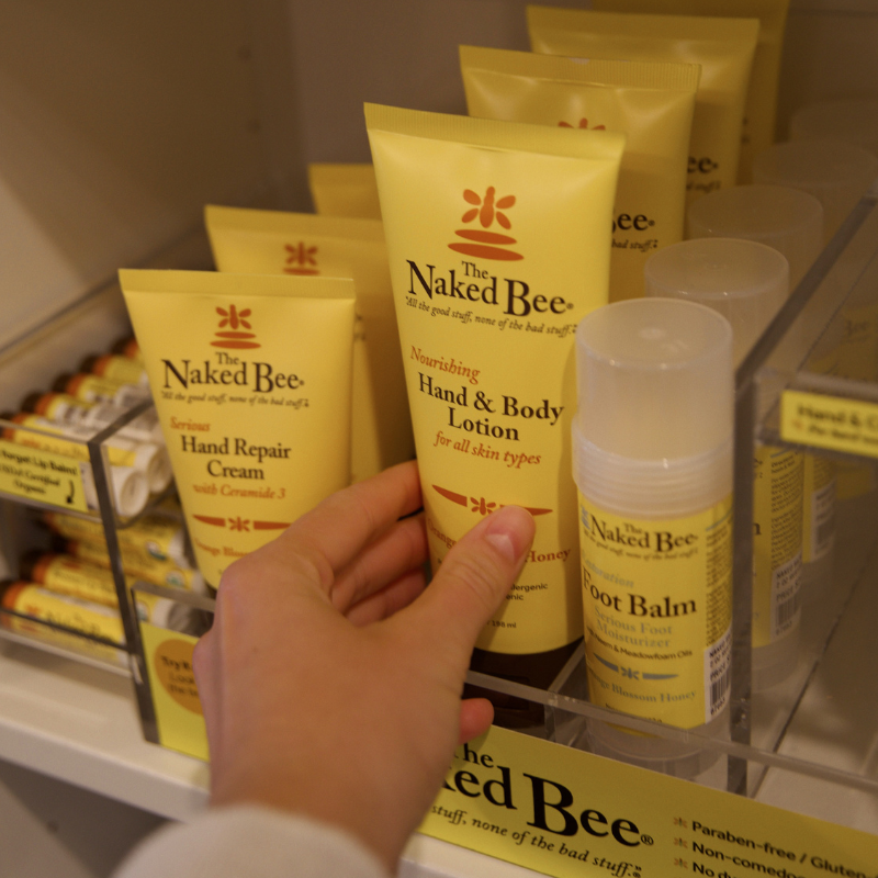 A hand holds a yellow tube of The Naked Bee Nourishing Hand & Body Lotion on a retail display shelf alongside other branded products.